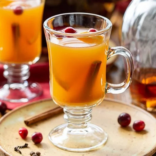 Apple cider in a tall glass with a handle. With cranberries floating as well as a submerged cinnamon stick